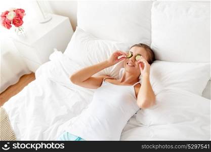 beauty, skin care and people concept - beautiful young woman lying in bed with cucumbers on face at home bedroom. beautiful woman applying cucumbers to face at home
