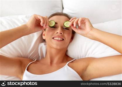 beauty, skin care and people concept - beautiful young woman lying in bed and applying cucumbers to her eyes at home bedroom. beautiful woman applying cucumbers to eyes at home