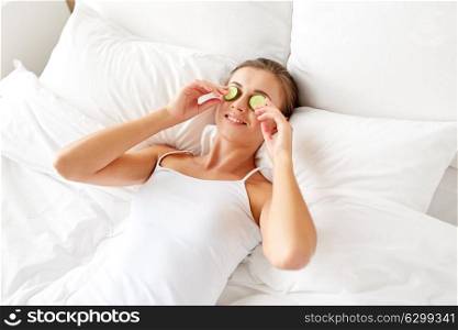 beauty, skin care and people concept - beautiful young woman lying in bed with cucumbers on face at home bedroom. beautiful woman applying cucumbers to face at home