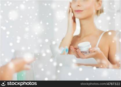 beauty, skin care and people concept - close up of smiling young woman applying cream to face mirror reflection at home bathroom over snow