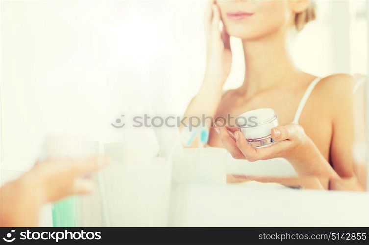 beauty, skin care and people concept - close up of smiling young woman applying cream to face mirror reflection at home bathroom. close up of woman applying face cream at bathroom