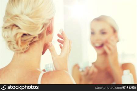 beauty, skin care and people concept - close up of smiling young woman applying cream to face and looking to mirror at home bathroom. close up of woman applying face cream at bathroom