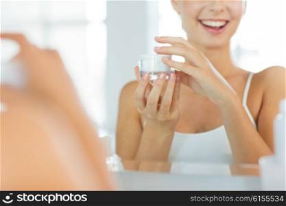 beauty, skin care and people concept - close up of smiling young woman with face cream mirror reflection at home bathroom