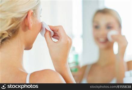 beauty, skin care and people concept - close up of smiling young woman cleaning her face with cotton disc and lotion at bathroom