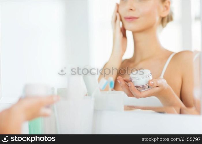beauty, skin care and people concept - close up of smiling young woman applying cream to face mirror reflection at home bathroom