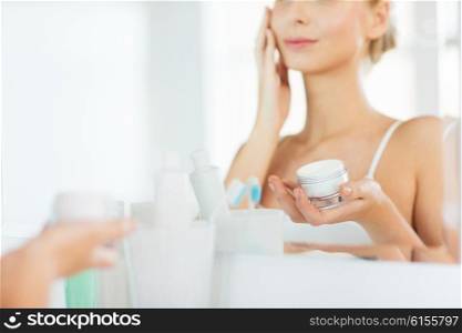beauty, skin care and people concept - close up of smiling young woman applying cream to face mirror reflection at home bathroom
