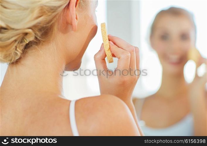 beauty, skin care and people concept - close up of smiling young woman washing her face with facial cleansing sponge at home bathroom