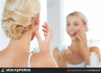 beauty, skin care and people concept - close up of smiling young woman applying cream to face and looking to mirror at home bathroom