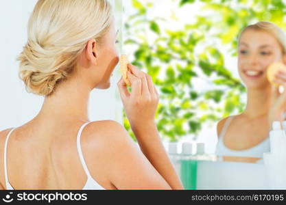 beauty, skin care and people concept - close up of smiling young woman washing her face with facial cleansing sponge at home bathroom over green natural background