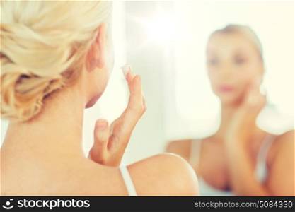 beauty, skin care and people concept - close up of young woman applying cream to face and looking to mirror at home bathroom. close up of woman applying face cream at bathroom. close up of woman applying face cream at bathroom