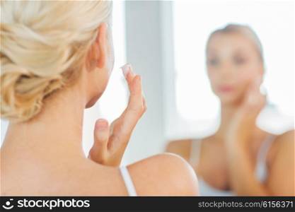 beauty, skin care and people concept - close up of young woman applying cream to face and looking to mirror at home bathroom