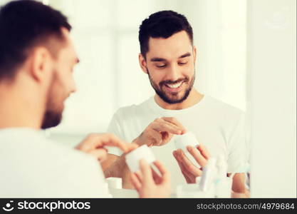 beauty, skin care and people concept - smiling young man applying cream to face and looking to mirror at home bathroom. happy young man applying cream to face at bathroom