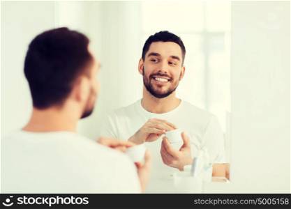 beauty, skin care and people concept - smiling young man applying cream to face and looking to mirror at home bathroom. happy young man applying cream to face at bathroom