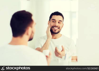 beauty, skin care and people concept - smiling young man applying cream to face and looking to mirror at home bathroom. happy young man applying cream to face at bathroom