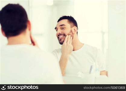 beauty, skin care and people concept - smiling young man applying cream to face and looking to mirror at home bathroom. happy young man applying cream to face at bathroom