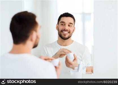 beauty, skin care and people concept - smiling young man applying cream to face and looking to mirror at home bathroom