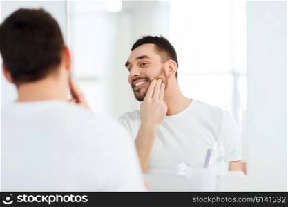 beauty, skin care and people concept - smiling young man applying cream to face and looking to mirror at home bathroom