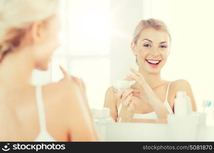 beauty, skin care and people concept - smiling young woman applying cream to face and looking to mirror at home bathroom. happy woman applying cream to face at bathroom