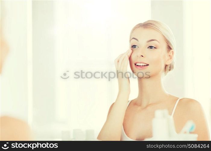 beauty, skin care and people concept - smiling young woman applying cream to face and looking to mirror at home bathroom. happy woman applying cream to face at bathroom. happy woman applying cream to face at bathroom