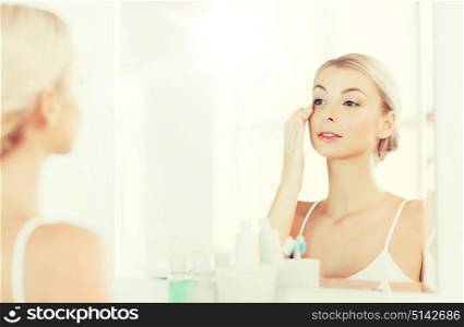 beauty, skin care and people concept - smiling young woman applying cream to face and looking to mirror at home bathroom. happy woman applying cream to face at bathroom