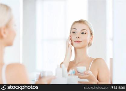 beauty, skin care and people concept - smiling young woman applying cream to face and looking to mirror at home bathroom