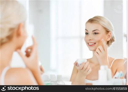 beauty, skin care and people concept - smiling young woman applying cream to face and looking to mirror at home bathroom