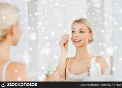 beauty, skin care and people concept - smiling young woman applying lotion to cotton disc for washing her face at bathroom over snow