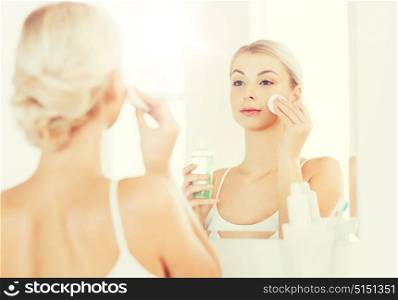 beauty, skin care and people concept - smiling young woman applying lotion to cotton disc for washing her face at bathroom. young woman with lotion washing face at bathroom