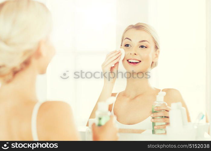 beauty, skin care and people concept - smiling young woman applying lotion to cotton disc for washing her face at bathroom. young woman with lotion washing face at bathroom