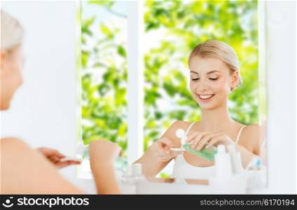 beauty, skin care and people concept - smiling young woman applying lotion to cotton disc for washing her face at bathroom over green natural background