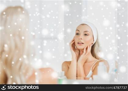 beauty, skin care and people concept - smiling young woman in hairband touching her face and looking to mirror at home bathroom over snow