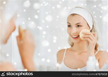beauty, skin care and people concept - smiling young woman in hairband washing her face with facial cleansing sponge at bathroom over snow