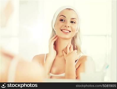 beauty, skin care and people concept - smiling young woman in hairband touching her face and looking to mirror at home bathroom. woman in hairband touching her face at bathroom
