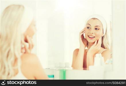 beauty, skin care and people concept - smiling young woman in hairband touching her face and looking to mirror at home bathroom. woman in hairband touching her face at bathroom. woman in hairband touching her face at bathroom