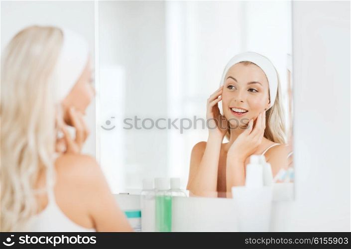 beauty, skin care and people concept - smiling young woman in hairband touching her face and looking to mirror at home bathroom
