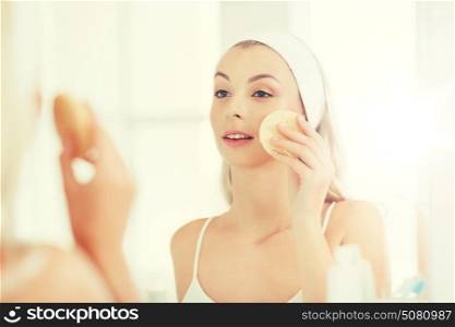 beauty, skin care and people concept - smiling young woman washing her face with facial cleansing sponge at bathroom. young woman washing face with sponge at bathroom