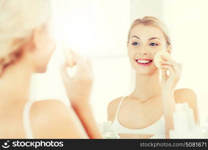 beauty, skin care and people concept - smiling young woman washing her face with facial cleansing sponge at bathroom. young woman washing face with sponge at bathroom