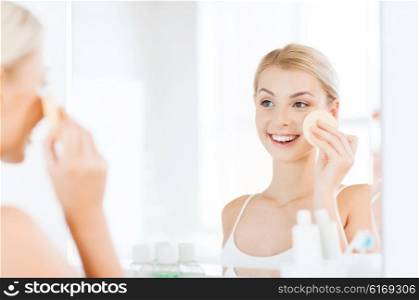 beauty, skin care and people concept - smiling young woman washing her face with facial cleansing sponge at bathroom