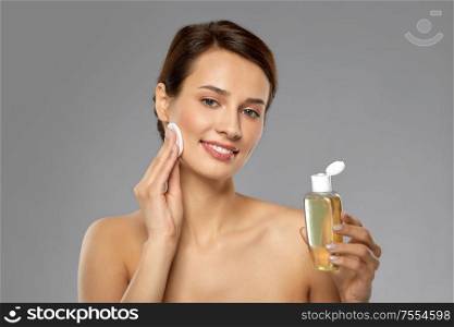 beauty, skin care and people concept - smiling young woman with toner or cleanser and cotton pad cleansing face over grey background. young woman with toner or cleanser and cotton pad