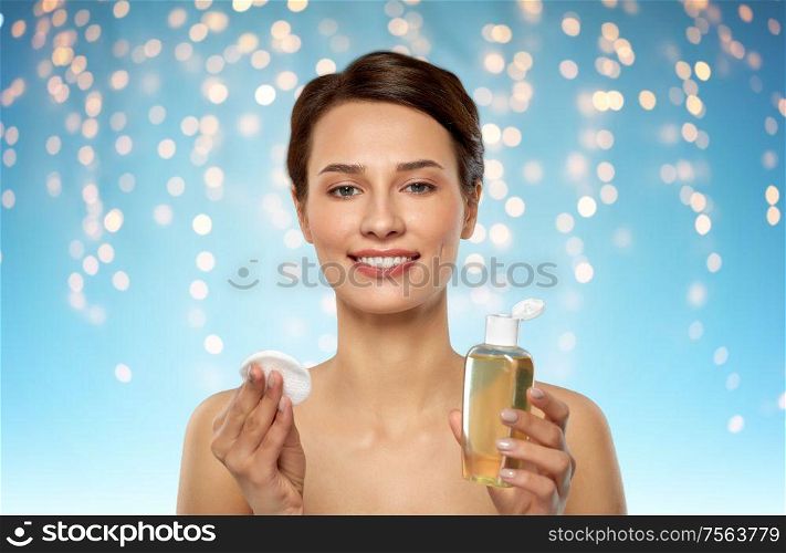 beauty, skin care and people concept - smiling young woman with toner or cleanser and cotton pad over holidays lights on blue background. young woman with toner or cleanser and cotton pad