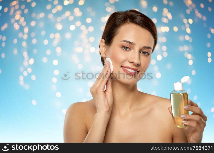 beauty, skin care and people concept - smiling young woman with toner or cleanser and cotton pad cleansing face over holidays lights on blue background. young woman with toner or cleanser and cotton pad