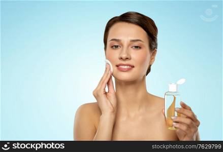 beauty, skin care and people concept - smiling young woman with toner or cleanser and cotton pad cleansing face over blue background. young woman with toner or cleanser and cotton pad