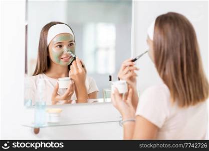 beauty, skin care and people concept - teenage girl with brush applying clay mask to face and looking in mirror at bathroom. teenage girl applying mask to face at bathroom