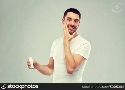 beauty, skin care, body care and people concept - smiling young man applying cream or lotion to face over gray background. happy young man applying cream or lotion to face