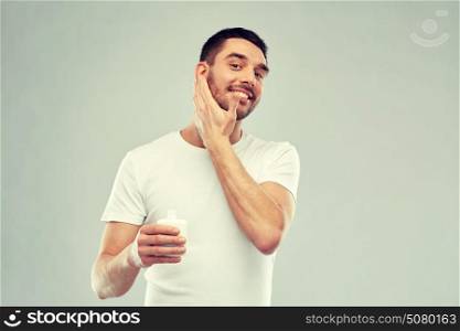 beauty, skin care, body care and people concept - smiling young man applying cream or lotion to face over gray background. happy young man applying cream or lotion to face