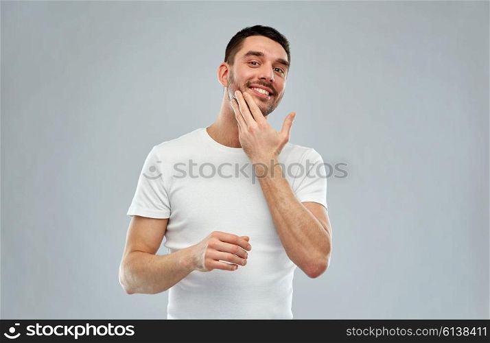 beauty, skin care, body care and people concept - smiling young man applying cream or lotion to face over gray background