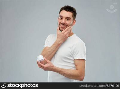 beauty, skin care, body care and people concept - smiling young man applying cream to face over gray background