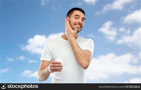 beauty, skin care, body care and people concept - smiling young man applying cream or lotion to face over blue sky and clouds background