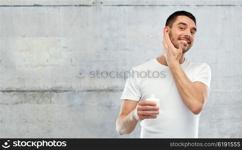 beauty, skin care, body care and people concept - smiling young man applying cream or lotion to face over gray wall background