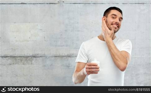 beauty, skin care, body care and people concept - smiling young man applying cream or lotion to face over gray wall background